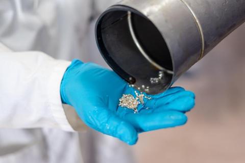 A gloved hand holds shiny metallic pieces being poured from a metal container,