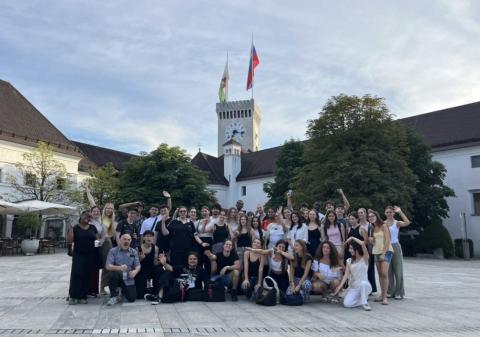 Group of around 40 young people in a town square, with old white buildings and a clock tower behind them   