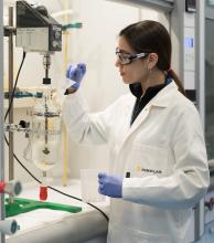 A woman in a white lab coat holding a small jug and a tool next to scientific equipment